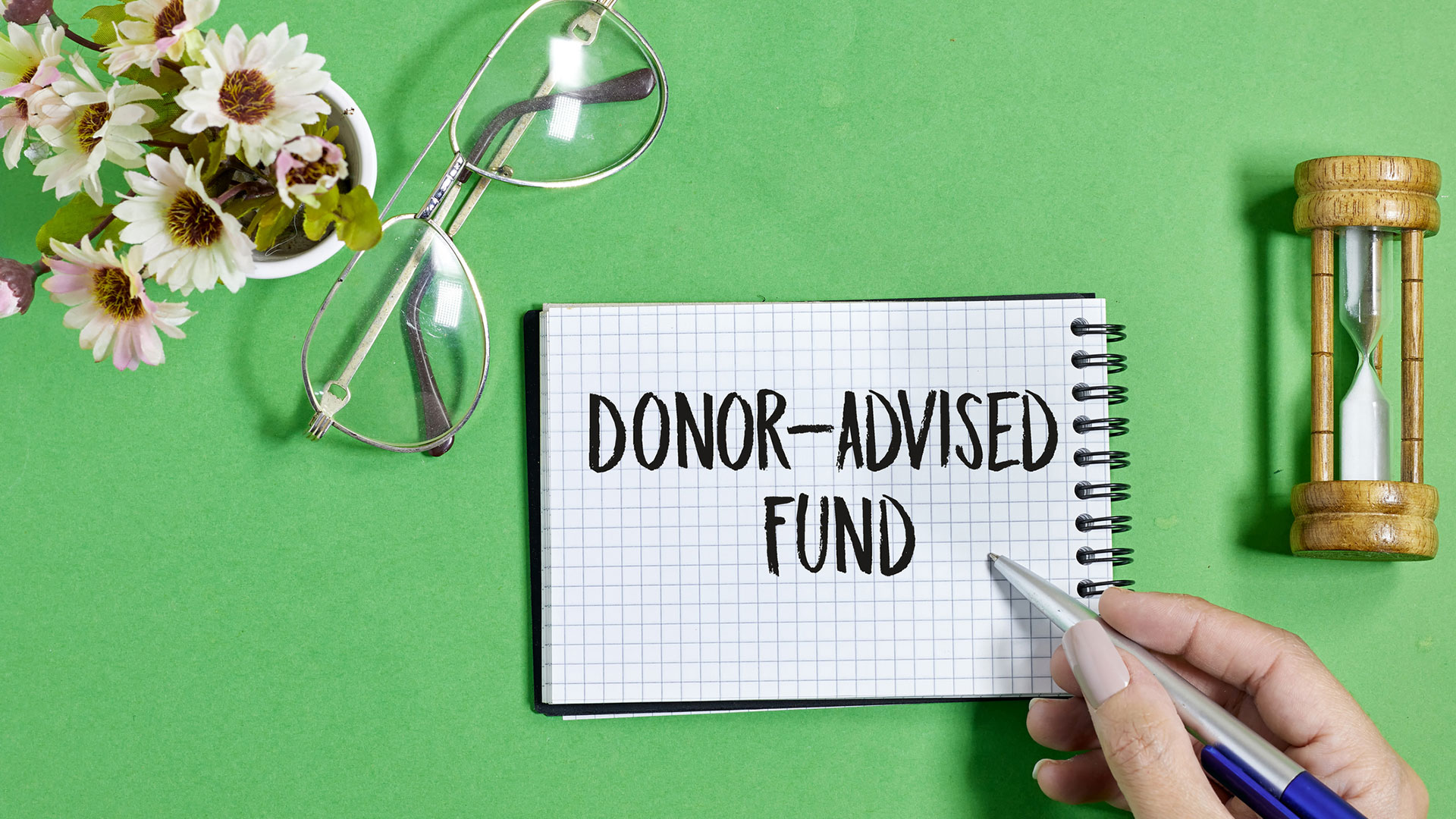 Donor-Advised-Funds - The INS Group Notebook page reads DONOR-ADVISED FUND as a hand writes with a pen nearby, with flowers, glasses, and an hourglass on a green background